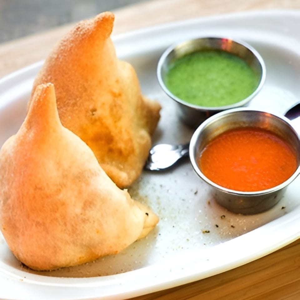 Two golden-brown samosas with green and red chutneys on a white plate at India Kitchen.